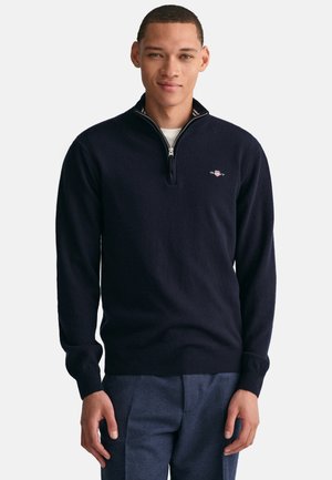 Navyblauwe zip-up trui gemaakt van zacht breimateriaal, met een hoge kraag en een klein logo op de borst. Geribde manchetten en zoom.