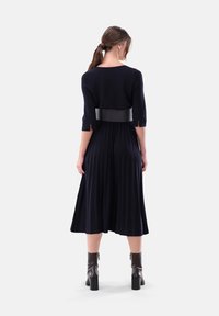 Navy gestricktes Kleid mit kurzen Ärmeln, versehen mit einem schwarzen Gürtel an der Taille und einem plissierten Rock. Grobe braune Stiefel mit Absatz runden den Look ab.