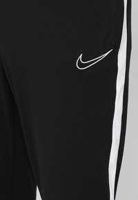 Schwarze Sporthosen mit weißen Seitenstreifen, aus glattem Stoff. Mit einem kleinen weißen Nike-Logo an der linken Hüfte.