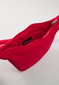 Borsa a tracolla rossa in tessuto con apertura con zip, dotata di una tasca interna. Texture liscia e design minimalista, contrassegnata con "PIECES."