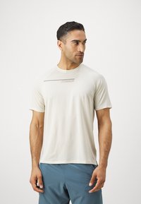 Light beige athletic t-shirt with short sleeves, featuring a crew neckline and a subtle logo design in a darker shade across the front.