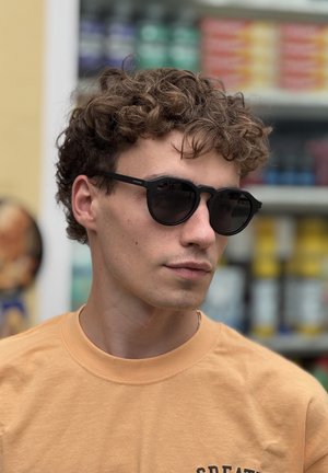 Young man with curly hair wearing black sunglasses and a light orange T-shirt, with colorful blurred shelves in the background.