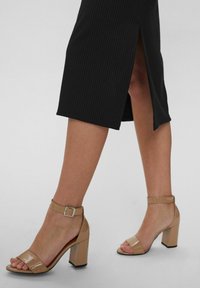 Beige High-Heelsandalen mit glänzender Oberfläche, offener Zehenpartie und Knöchelriemen. Getragen mit einem schwarzen, gerippten Midikleid mit seitlichem Schlitz.