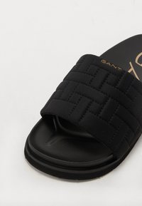 Sandală neagră tip slip-on, cu un upper din țesătură împletită, talpă interioară texturată și talpă exterioară din cauciuc neted. Prezintă un logo auriu pe curea.