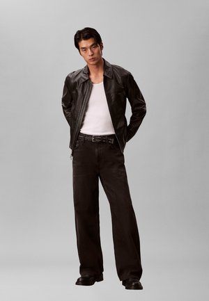 Junger Mann trägt schwarze Lederjacke, weißes Tanktop, schwarze weite Jeans mit Nietengürtel und schwarze Schuhe, steht mit den Händen hinter dem Rücken.