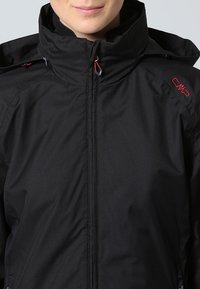 Personne portant une veste noire zippée avec un col montant et un logo rouge sur l'épaule.