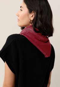 Red textured scarf draped around the neck, paired with a black knitted top. Scarf features soft, frayed edges and a subtle sheen.