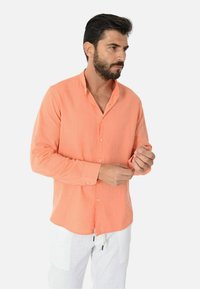 Camicia di lino arancione chiaro con colletto a bottoni, maniche lunghe e bottoni lungo la parte anteriore, indossata con pantaloni bianchi.