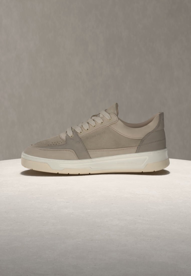 Beige and gray low-top sneaker with white sole and laces, displayed on a light round surface against a neutral background.