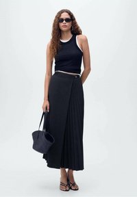 Top noir côtelé avec bord blanc, associé à une longue jupe plissée noire et des sandales noires. Tenant un sac à main en osier noir.