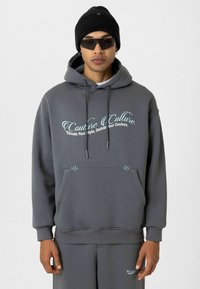 Grijze hoodie van zacht materiaal, met een voorzak, gegolfde naden en een teksten ontwerp in lichtblauw. Gecombineerd met grijze onderkleding.