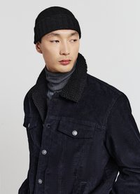 Junger Mann mit schwarzer Strickmütze, dunkler Cordjacke mit Fleecekragen und grauem Rollkragenpullover, der seitlich vor einem schlichten Hintergrund blickt.