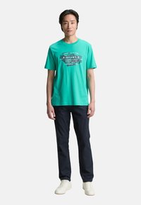 Turkis bomuld t-shirt med et grafisk tryk i sort og hvid, parret med marineblå bukser og hvide sneakers. Enkel rund halsudskæring.