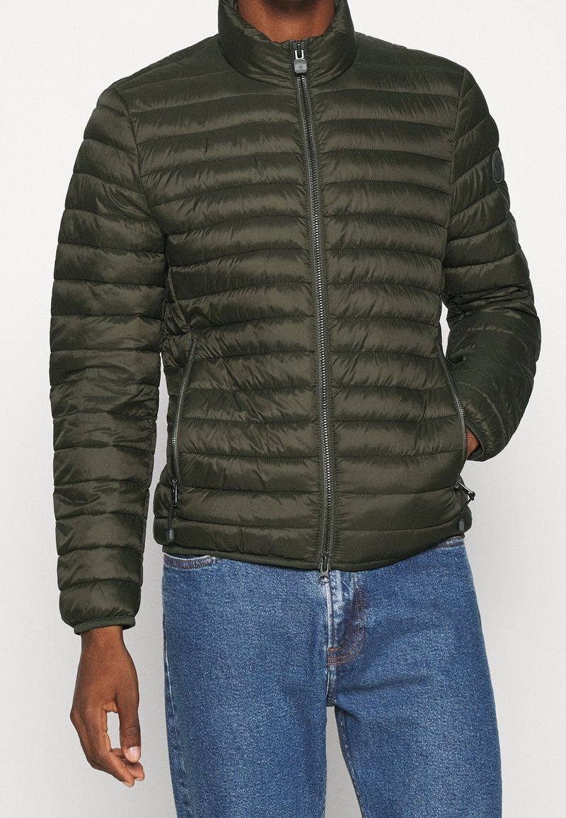 Olivgrüne gesteppte Jacke mit hohem Kragen, Reißverschluss und Seitentaschen. Mit horizontalen Steppnähten und einem Logopatch am Ärmel versehen.