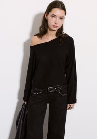 Črni pulover brez naramnic z dolgimi rokavi, v kombinaciji z visokim pasom črnimi kavbojkami, ki imajo okrasne kovinske detajle in črn pas.