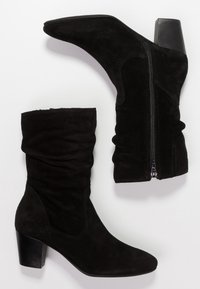 Une paire de bottes en suède noir à mi-mollet avec des talons bloc et des fermetures éclair sur le côté, une botte debout et l'autre à plat sur un fond blanc.