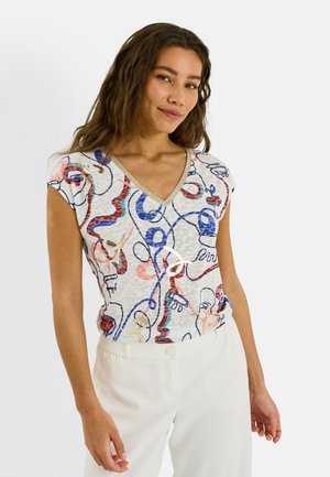 Femme portant un haut blanc à col V avec des motifs abstraits colorés en lignes et un pantalon blanc, debout devant un fond uni.