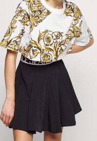 Zkrácená bílá tričko s ozdobnými zlatými vzory, kombinované s rozšířenou černou sukní. Tričko má kontrastní tištěný pruh.