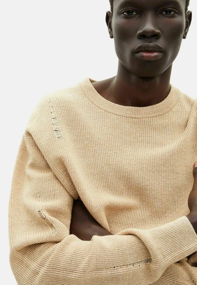Beige gestrickter Pullover mit runder Ausschnitt, ausgestattet mit gerippten Bündchen und einem leicht strapazierten Detail an der Schulter. Weicher, strukturierten Stoff.
