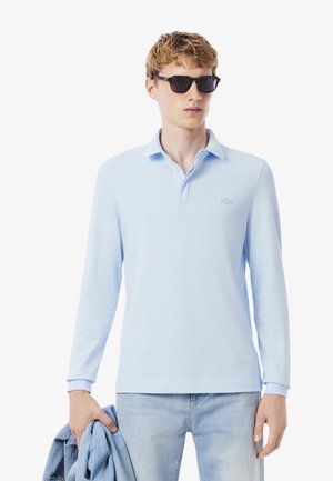 Joven con una camiseta de polo de manga larga azul claro, gafas de sol, sosteniendo una chaqueta de mezclilla, de pie contra un fondo blanco.