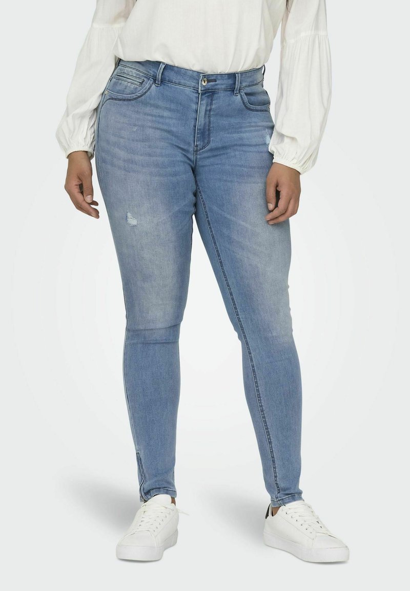 Svetlomodré úzke džínsy s miernym opotrebovaním na kolene; obsahujú predné vrecká, zips a sú spárované s bielymi teniskami.