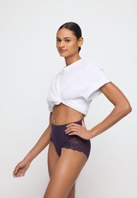 Hoge paarse slip met kantdetails in een contrasterende donkere tint, gecombineerd met een geknoopt wit T-shirt met opgerolde mouwen.