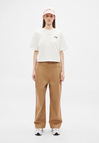 Camiseta blanca recortada con logo verde, pantalones marrones de pierna ancha y gorra rosa con texto. Combinado con zapatillas deportivas blancas y grises.