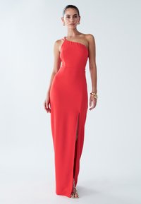 Punainen yhden olkapään iltapuku, jossa on reiden korkuinen halkio. Valmistettu sileästä kankaasta, istuva malli hienovaraisilla olkapääyksityiskohdilla.