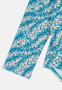 Tkano v teali barvi z cvetličnim vzorcem, ki vključuje majhne bele in rožnate cvetove, okrašeno z svetlo modrimi mašnicami. Gladka tekstura, lahka zasnova.