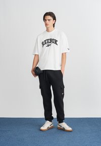 Balta medvilninė marškinėliai su juodu "Reebok" logotipu, derinama su juodais krovinių kelnėmis ir kreminiais sportbačiais su juodais akcentais bei guminiu padu.