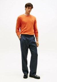 Oranges Rundhals-Sweatshirt, kombiniert mit dunkelblauen, gefalteten Hosen. Schwarze Schuhe runden das Outfit ab. Einfaches, sauberes Design und maßgeschneiderte Passform.