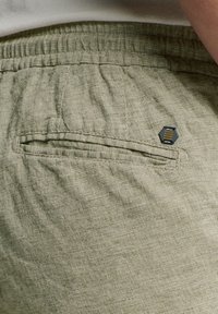 Lichtgroene linnen shorts met een gestructureerd oppervlak, voorzien van een achterzak en een klein hexagonaal merklabel in gedempte kleuren.