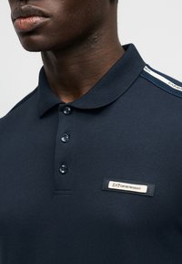 Μπλούζα πόλο σε ναυτικό μπλε χρώμα, κατασκευασμένη από απαλό βαμβάκι με κουμπωτό πατιλέτα, που διαθέτει υφασμάτινη λεπτομέρεια στον ώμο και ετικέτα με το λογότυπο της εταιρείας.