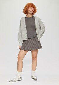Cardigan en tricot gris clair avec de gros boutons, porté sur une chemise gris foncé et une jupe plissée grise à rayures blanches, accompagnée de chaussures argentées.