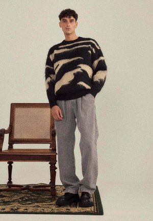 Pull en laine rayé noir et crème, coupe décontractée ; pantalon plissé gris ; chaussures noires épaisses ; chaise en bois et tapis à motifs en arrière-plan.