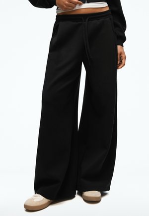 Personne portant un pantalon noir à jambes larges avec une taille à cordon et des baskets beiges, debout devant un fond blanc uni.