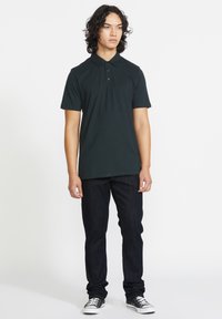 Polo shirt noir en coton avec un col classique et une patte de boutonnage à trois boutons, associé à un jean foncé et des baskets noires.
