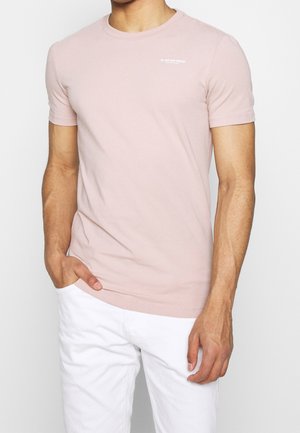 Mann trägt hellrosa kurzärmeliges T-Shirt mit kleinem Logo auf der Brust und weiße Hose, Hand in der Tasche, vor hellem einfarbigem Hintergrund.