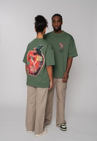 Deux personnes portant des t-shirts graphiques verts assortis et des pantalons beiges se tiennent devant un fond uni ; l'une est de face, l'autre montre un grand motif de pomme.