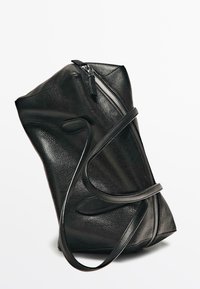 Sac fourre-tout en cuir noir texturé avec des poignées longues et une fermeture éclair argentée visible sur un côté, présenté sur un fond blanc uni.