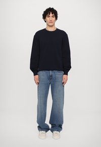 Sweatshirt azul-marinho com decote redondo, mangas compridas e um detalhe texturizado. Combinado com calças de ganga de perna larga azul claro e ténis brancos.