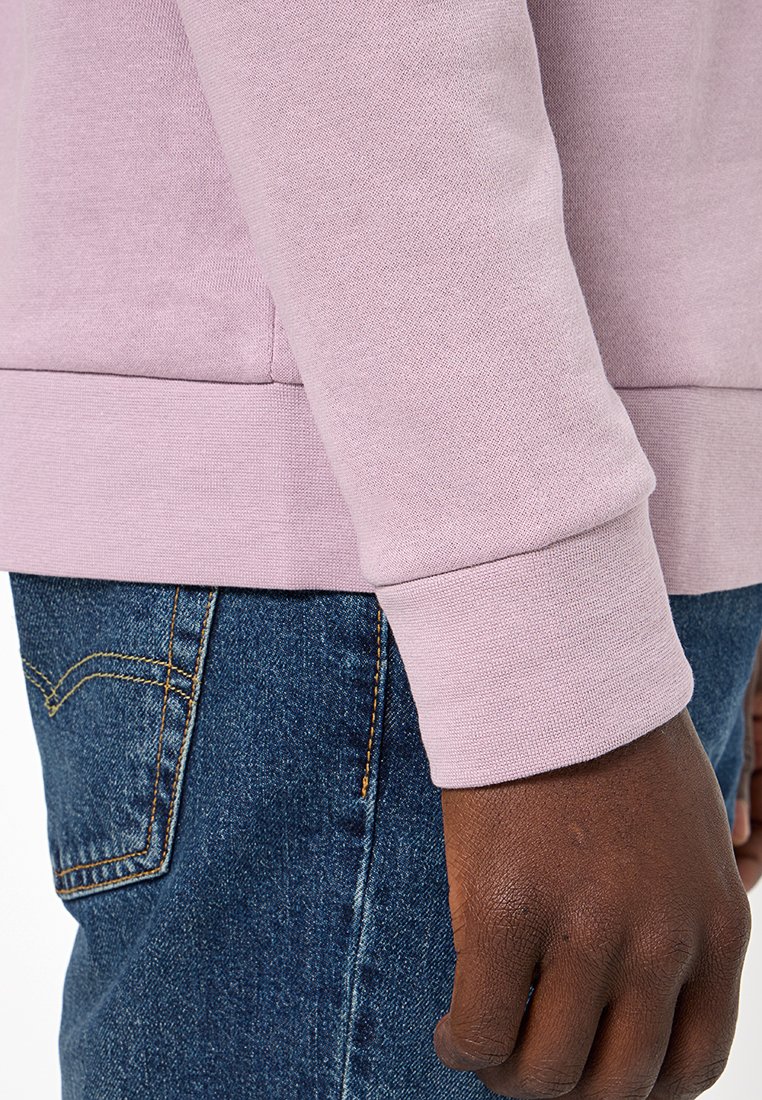 Una mano di carnagione scura appoggiata accanto alla tasca dei jeans blu, che indossa una maglietta a maniche lunghe di colore rosa chiaro con polsini e vita a coste.
