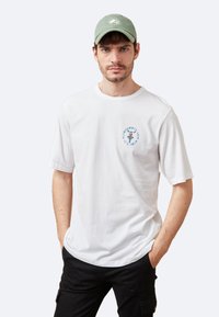 Camiseta de algodón blanca de manga corta, con un gráfico circular azul en el pecho. Combinada con una gorra verde claro. Ajuste casual.