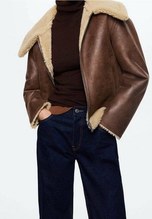 Veste en shearling marron avec doublure en polaire crème, portée sur un pull à col roulé marron foncé et un jean bleu, les mains dans les poches.