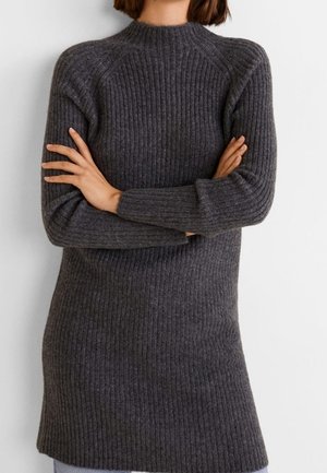 Personne portant un pull long en maille côtelée gris foncé avec des manches longues et un col montant, les bras croisés sur la poitrine.
