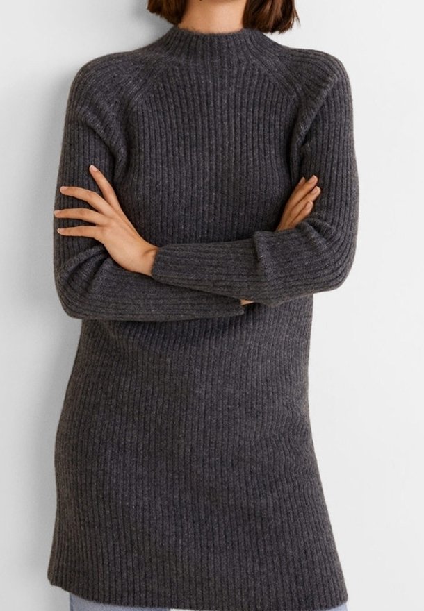 Personne portant un pull long en maille côtelée gris foncé avec des manches longues et un col montant, les bras croisés sur la poitrine.