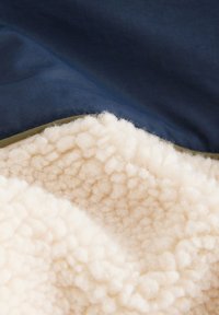 Tela de forro polar texturizada en crema, superpuesta con tela azul marino y ribete verde oliva, mostrando una textura suave y esponjosa y colores contrastantes.