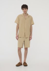 Camicia a maniche corte color sabbia e pantaloni corti abbinati in un tessuto testurizzato, con bottoni davanti. Indossati con scarpe scivolate marroni.