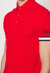 Rød polo t-shirt lavet af tekstureret stof, med en treknaps-lukning, hvide og marineblå stribede detaljer på ærmerne og broderet logo.