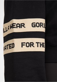 Sudadera negra con una banda ancha color crema y texto negro que dice "Gorilla Wear For The Motivated" a lo largo de la manga.